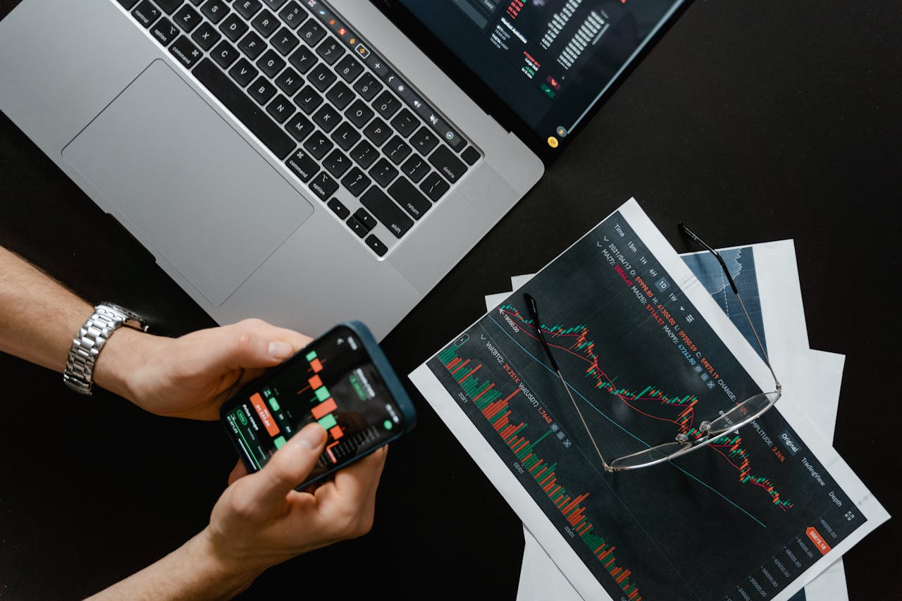 About Hands using smartphone beside laptop with stock charts, showcasing digital trading.